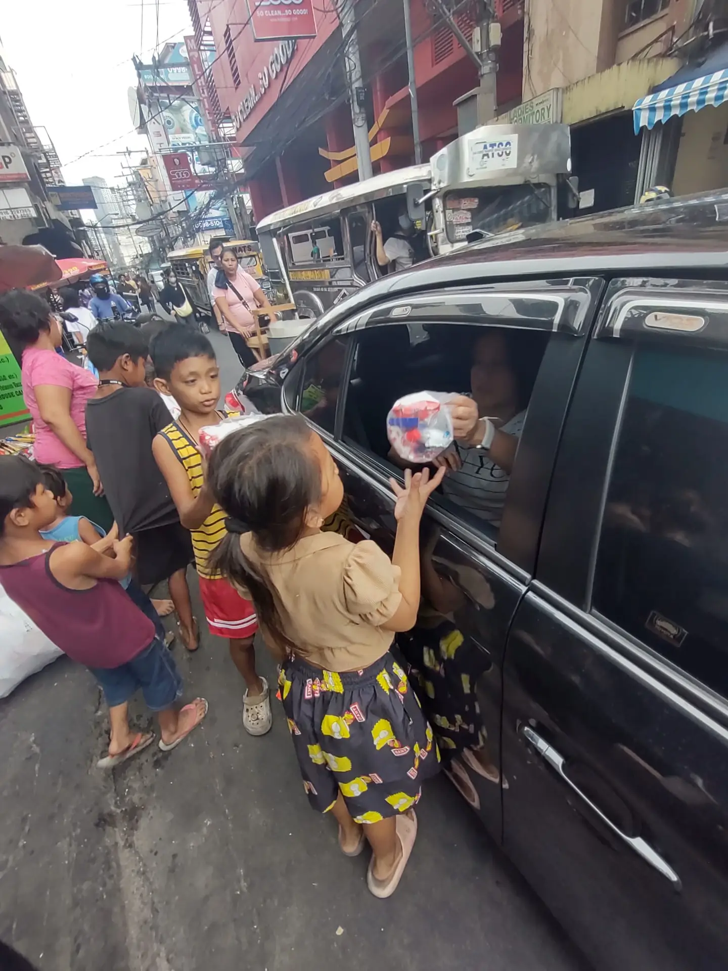 Children on a busy Manila street reaching for food packs handed through a car window
